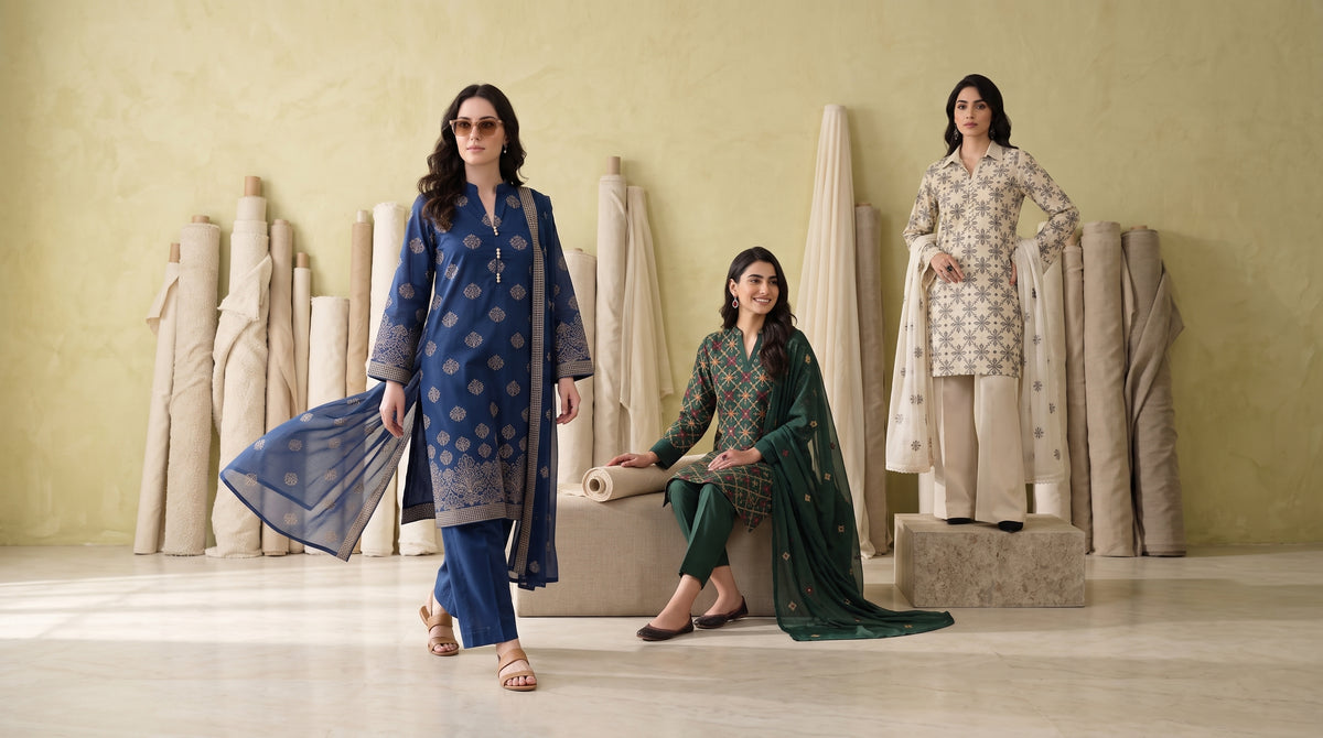 Three women modeling traditional South Asian salwar kameez suits in a studio setting with fabric rolls in the background. The suits include a blue patterned kameez with matching pants and dupatta, a dark green patterned kameez with matching pants and dupatta, and a beige patterned kameez with matching pants and dupatta.