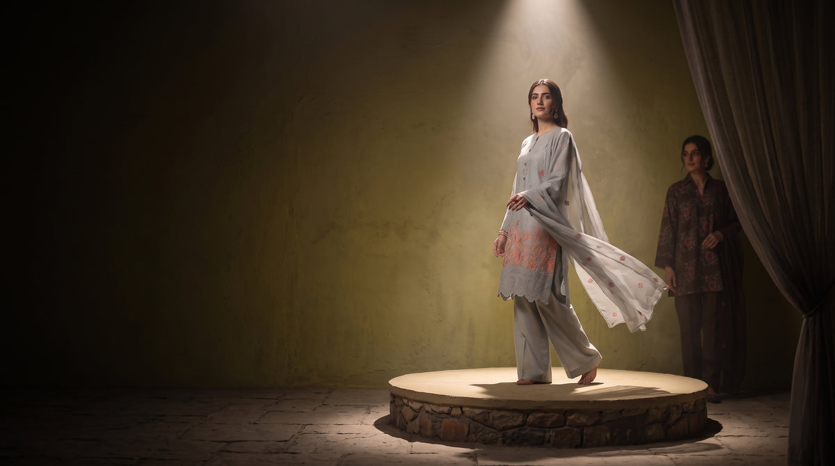 A model in a light gray and pink embroidered traditional outfit stands on a circular stone platform under a spotlight, with another person standing in the background near a curtain.