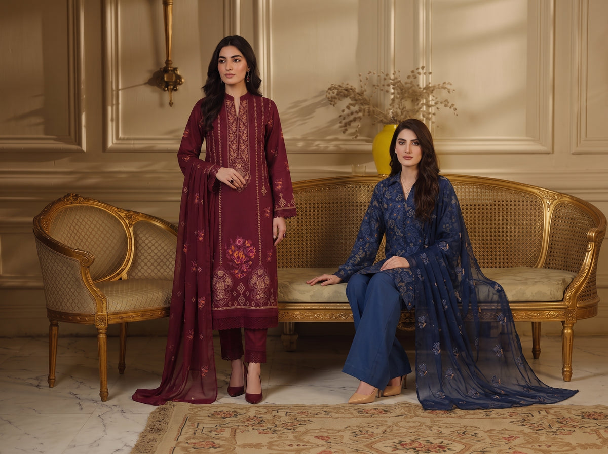 Two women modeling traditional South Asian salwar kameez suits in an elegant room. The woman standing wears a deep maroon/burgundy suit with gold embroidery, and the woman seated wears a navy blue suit with floral embroidery.